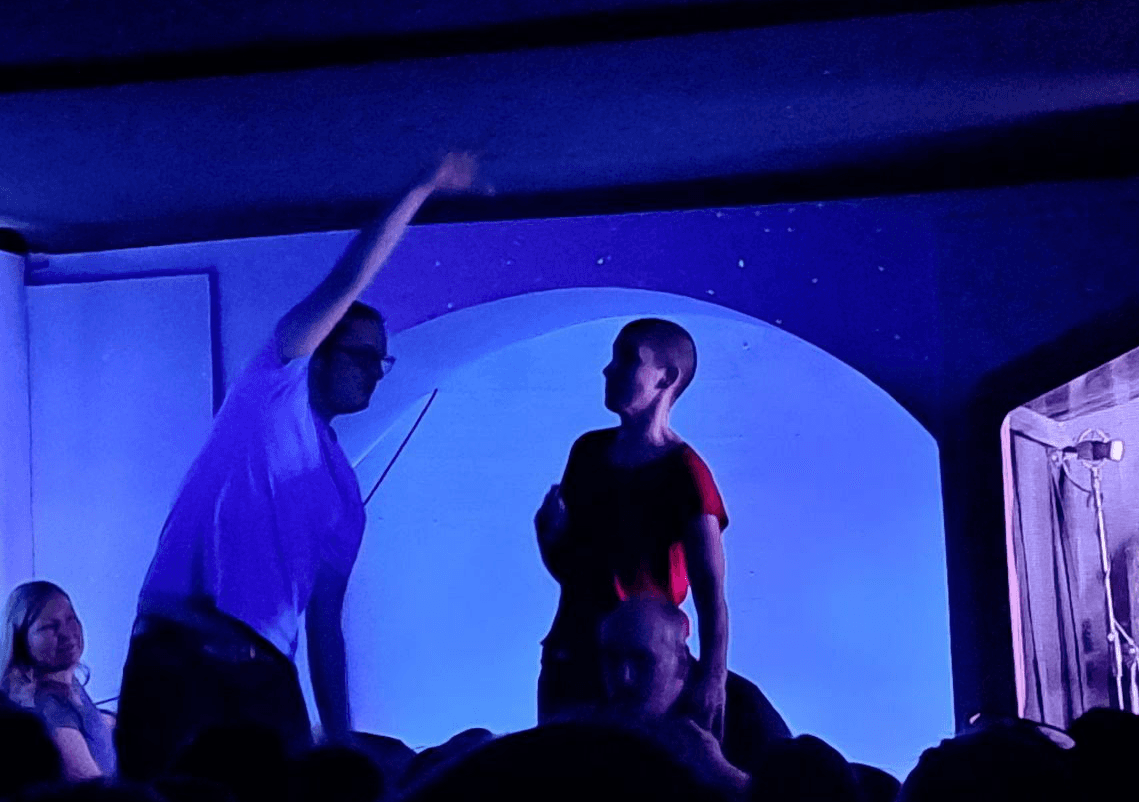 Zwei Schauspieler auf der Bühne eines Improtheaters, beleuchtet von blauem Licht. Eine Person in einem weißen T-Shirt hebt den Arm, während die andere Person, im roten T-Shirt, sie beobachtet.