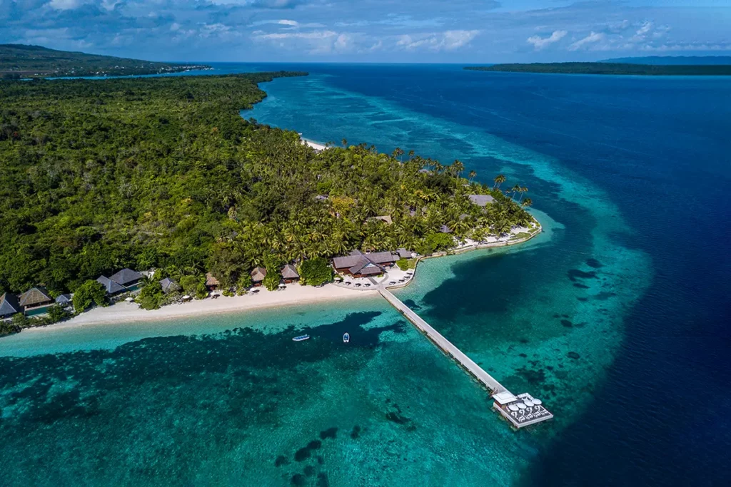 Weite Luftaufnahme eines Inselresorts mit einem sehr langen weißen Steg, der weit in das korallenreiche, klare Wasser ragt, umgeben von dichtem Palmenbewuchs und kleinen Villen am Strand.