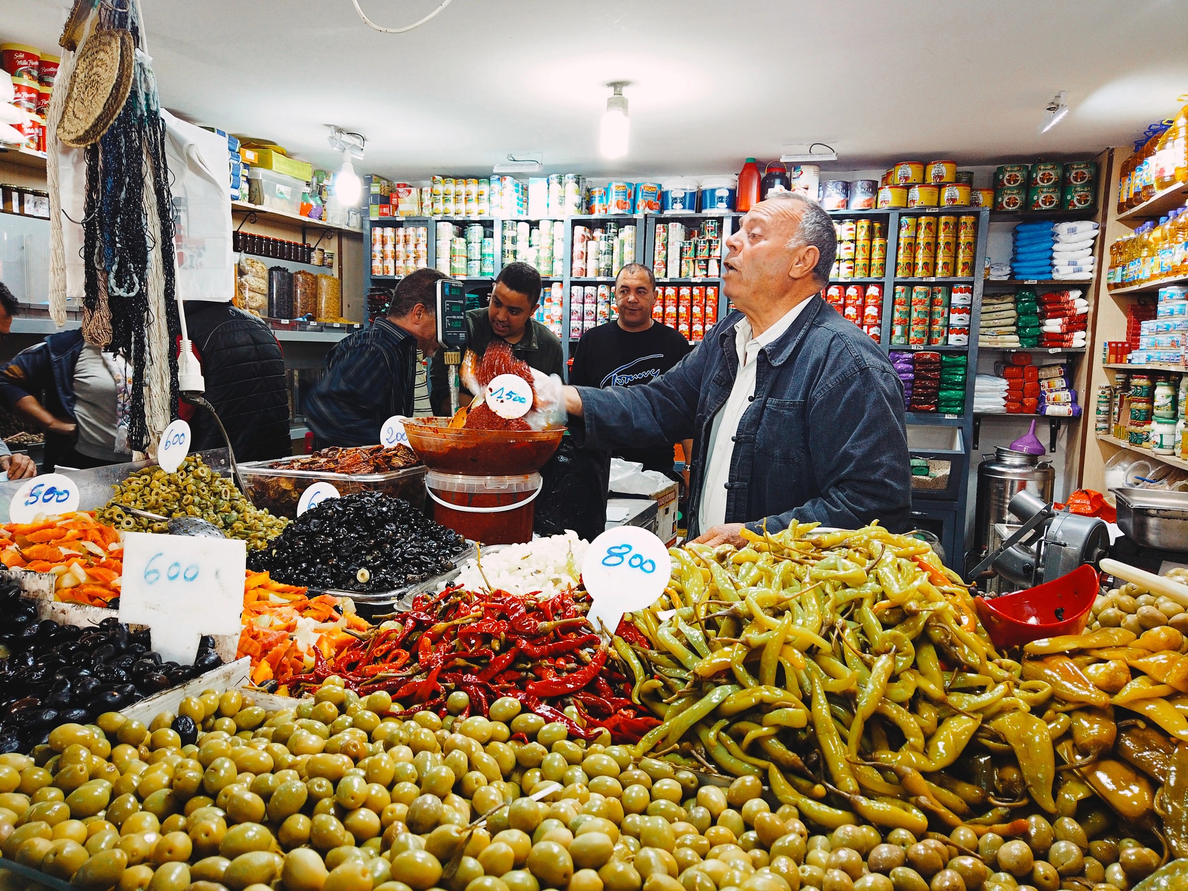 Photographie documentaire d'un marché à Tunis montrant un marchand entouré par ses produits et lgéumes tendre un bol d'épices à son client pendant que ses amis discutent en arrière plan