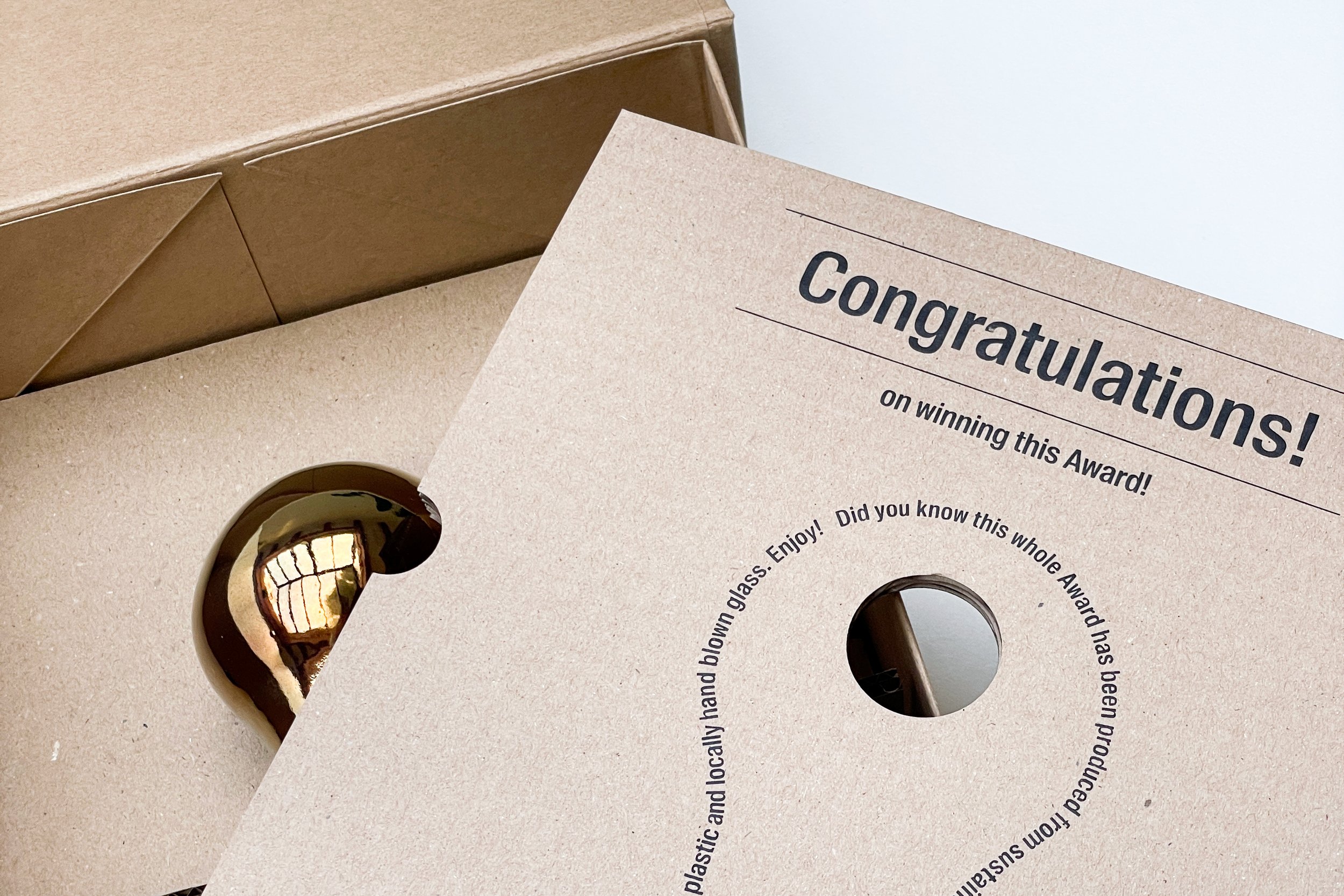 Image of the golden glass award in an untreated cardboard box with cardboard inlay with laser-engraved text