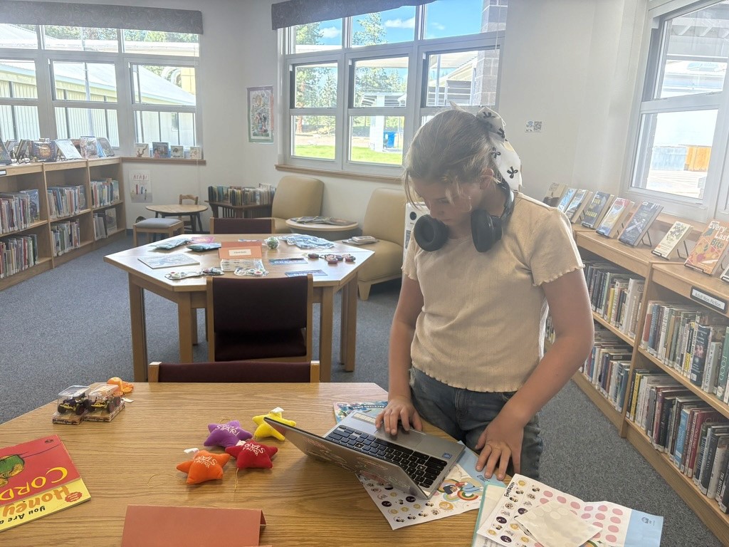 A young person using a laptop at a library table, with books and activity materials nearby.