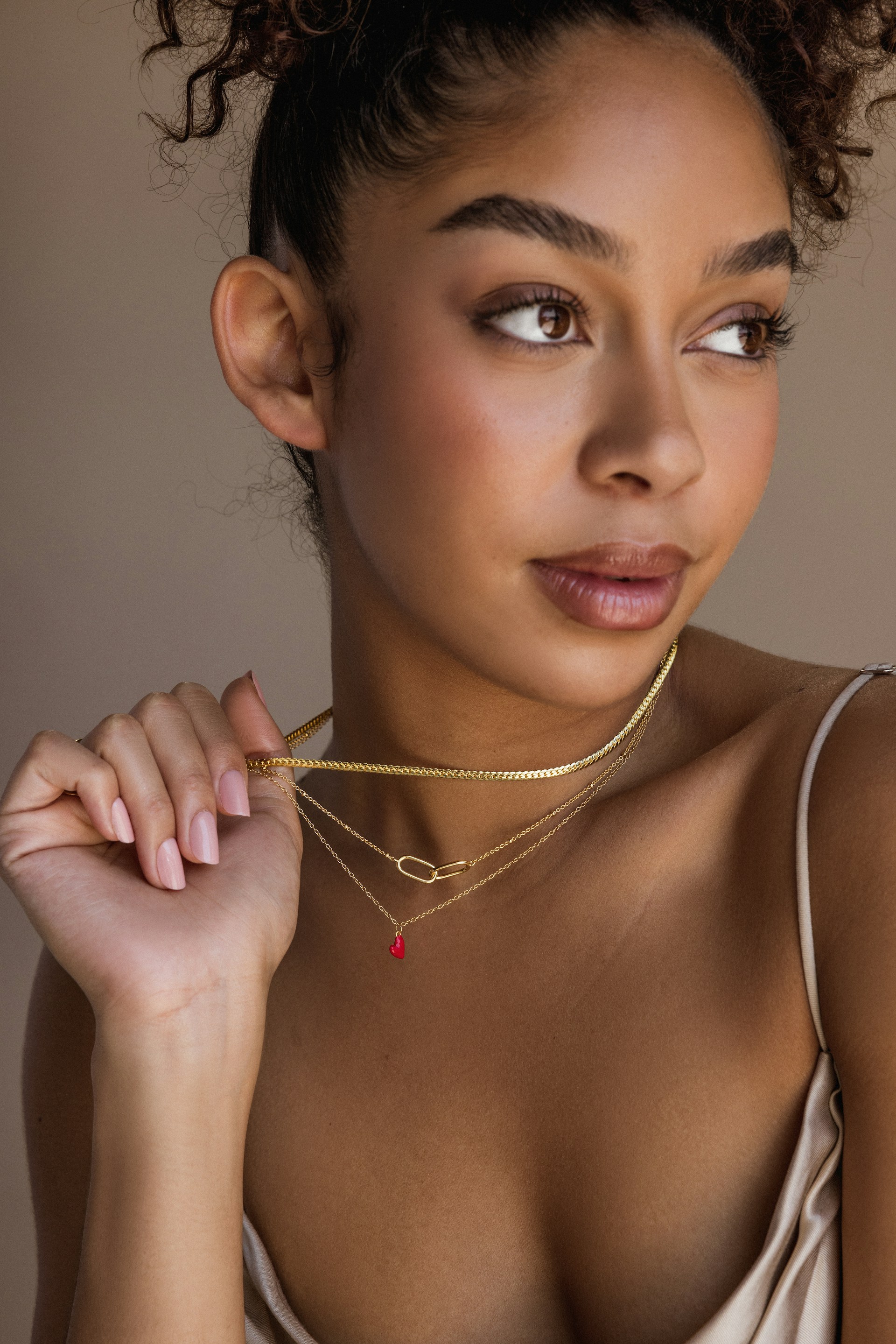 Portrait of a woman with a curly updo looking off-camera, gently holding her layered gold necklaces.