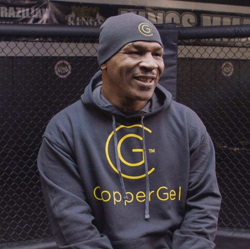 Mike Tyson wearing a black hoodie with a logo smiles while sitting in front of a building.