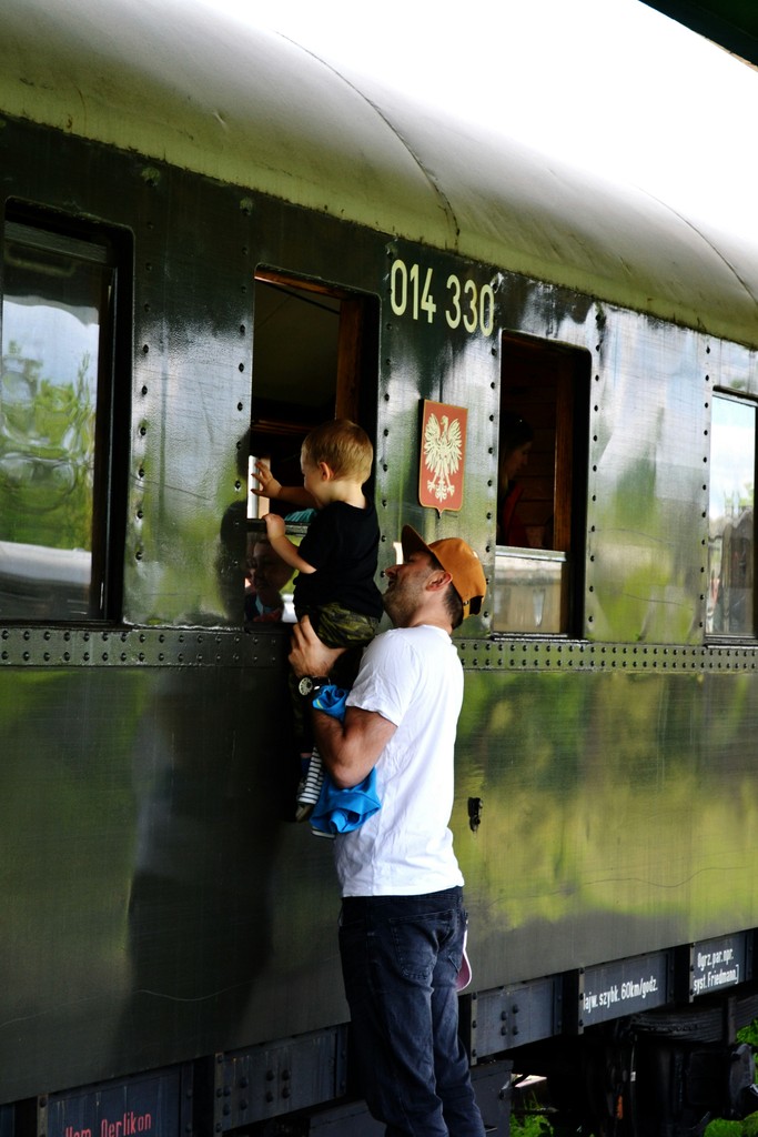 A father lifts his young son to look through the window of an old train carriage, marked with the number 014 330 and a crest, evoking a sense of adventure and exploration during a family outing.