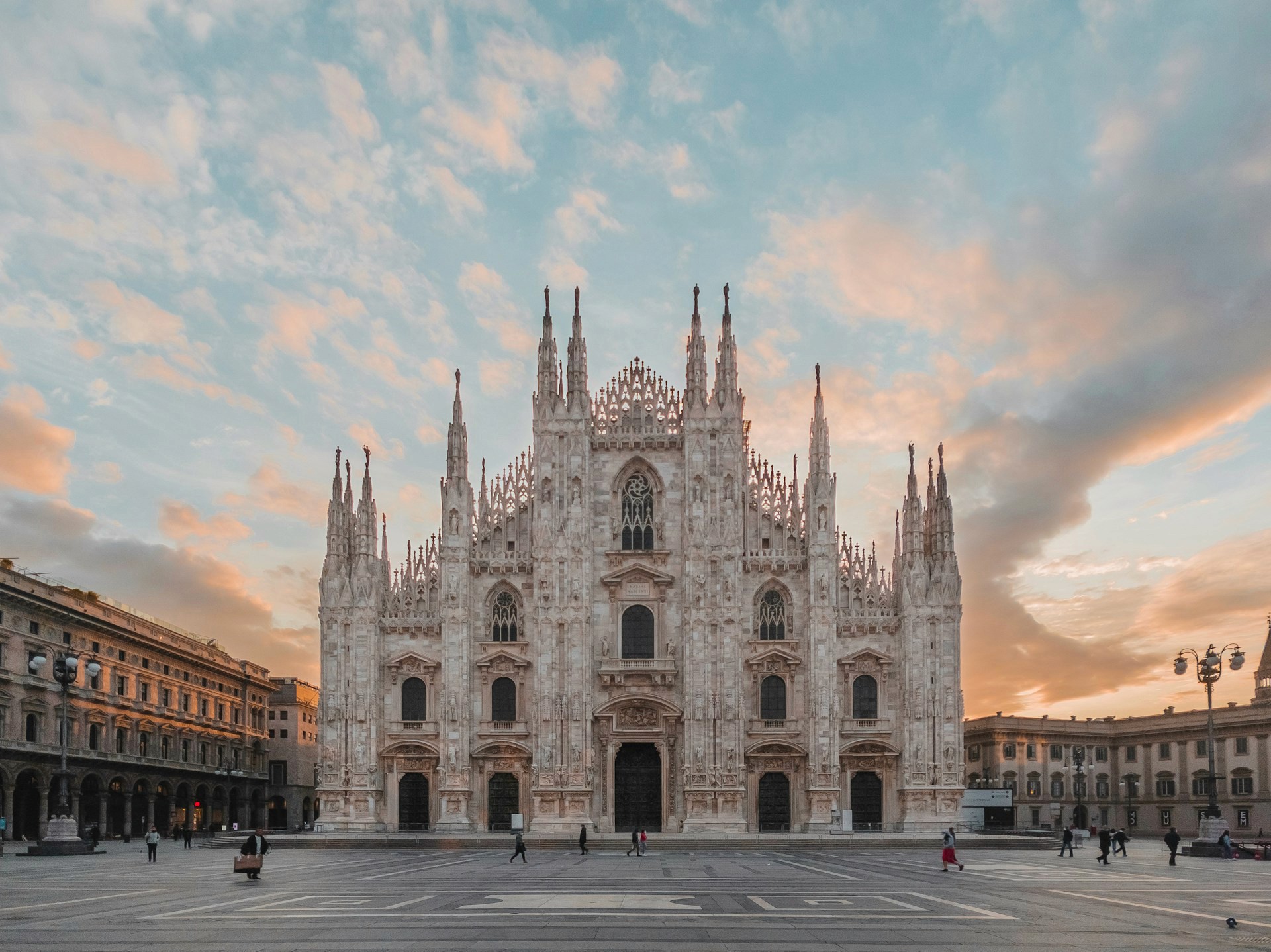A wide shot of the Milan Cathedral (Duomo di Milano) at sunset, its Gothic spires silhouetted against a soft blue and orange sky over a large, open plaza.