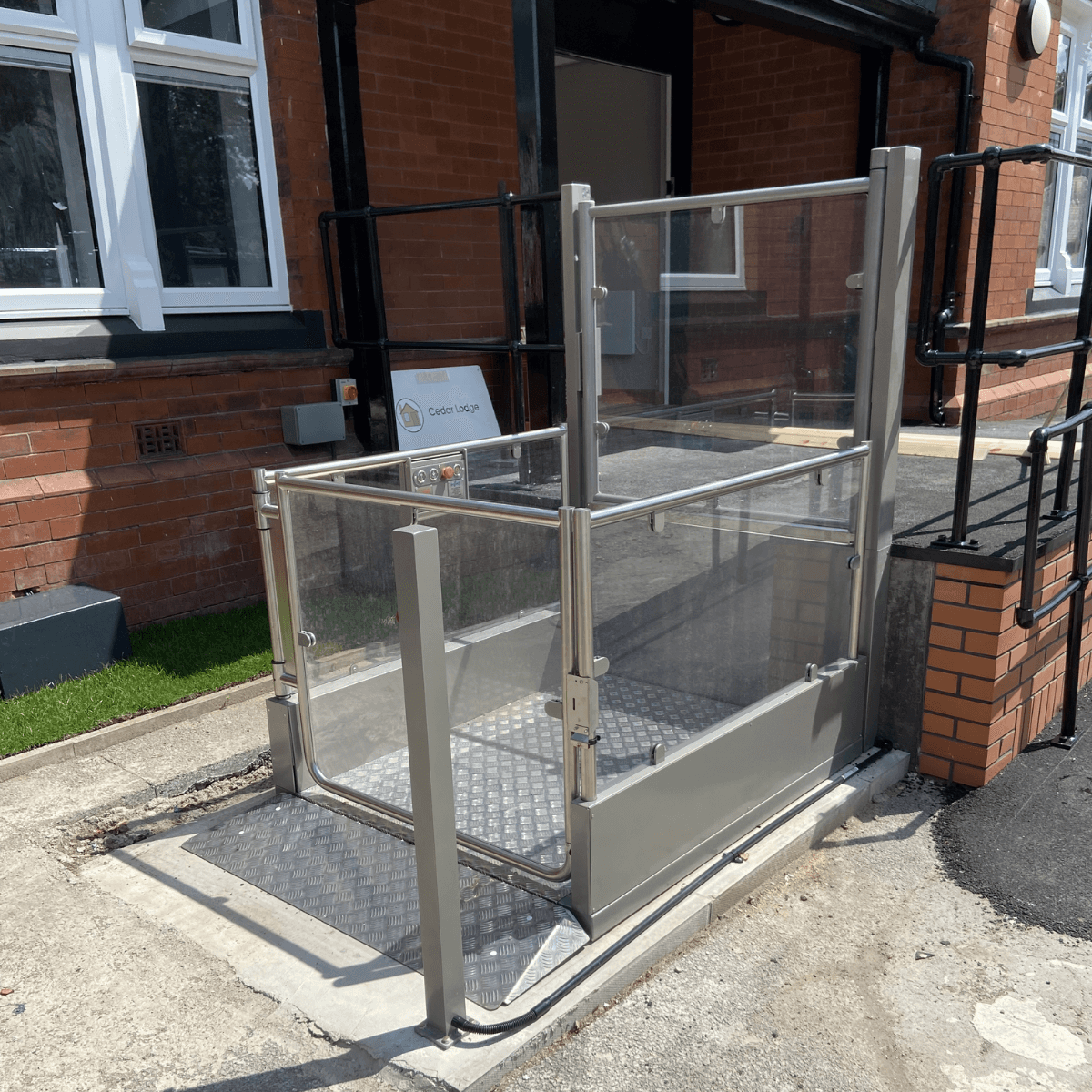 An outdoor vertical platform lift with glazed side panels installed at the entrance of a UK care home, providing wheelchair access to a raised threshold with a hinged gate and ground-level controls.