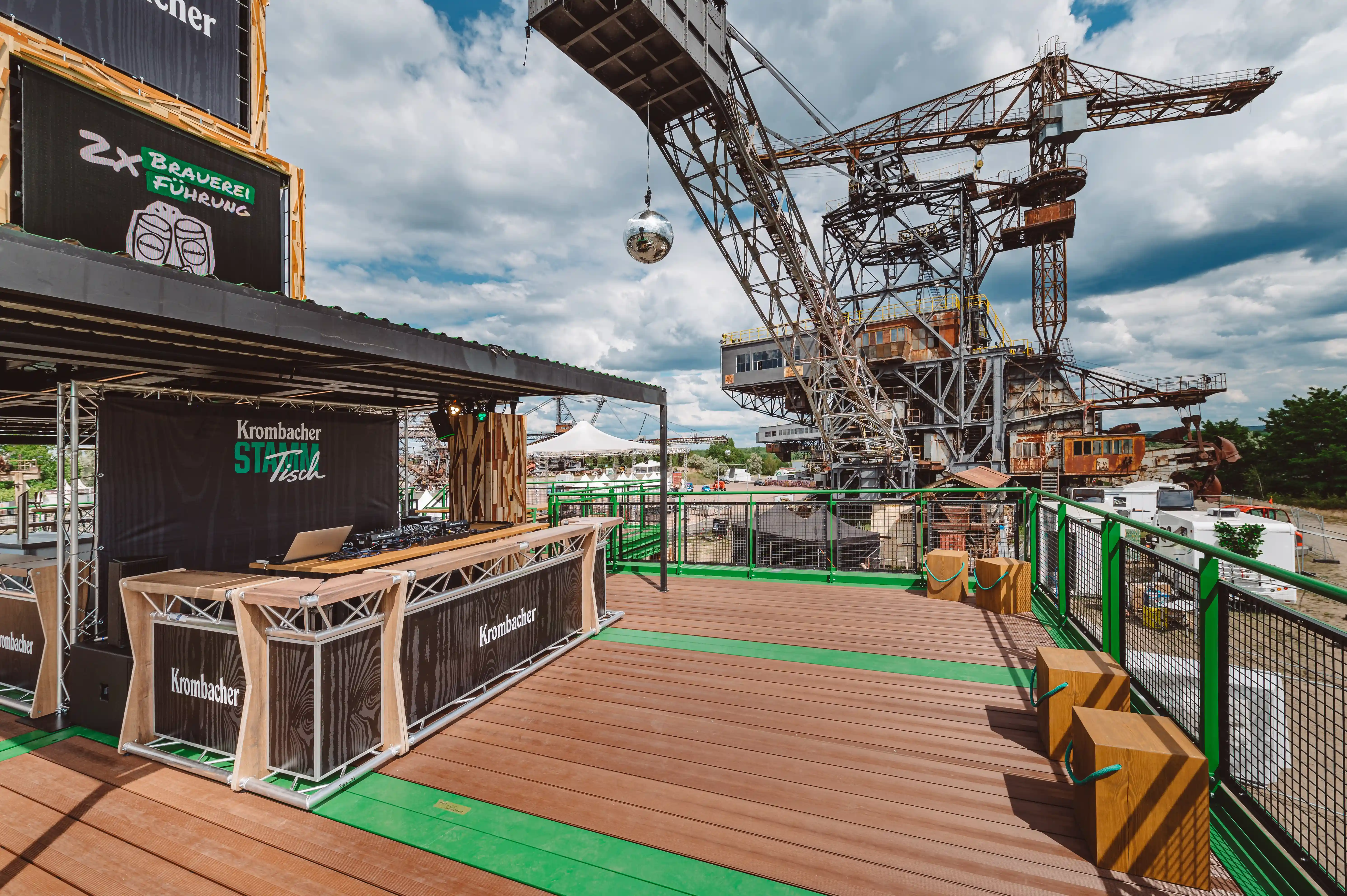 Bar terrace of the KROMBACHER festival stand at Melt with wooden deck, branded bar counters, DJ desk and the rusty industrial cranes of Ferropolis behind.