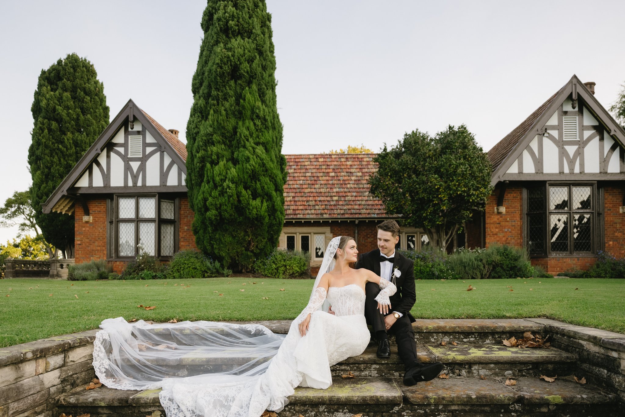 Bride and groom seated on stone steps with veil trailing, in front of Tudor-style houses and gardens