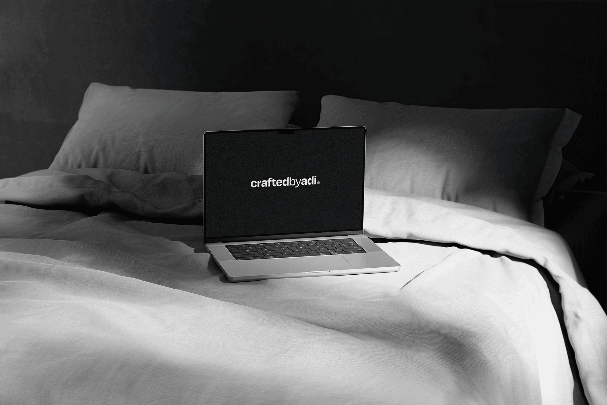 A silver laptop open on a bed with rumpled grey linens, displaying the 'craftedbyadi' logo on a dark screen under soft, dramatic window lighting.
