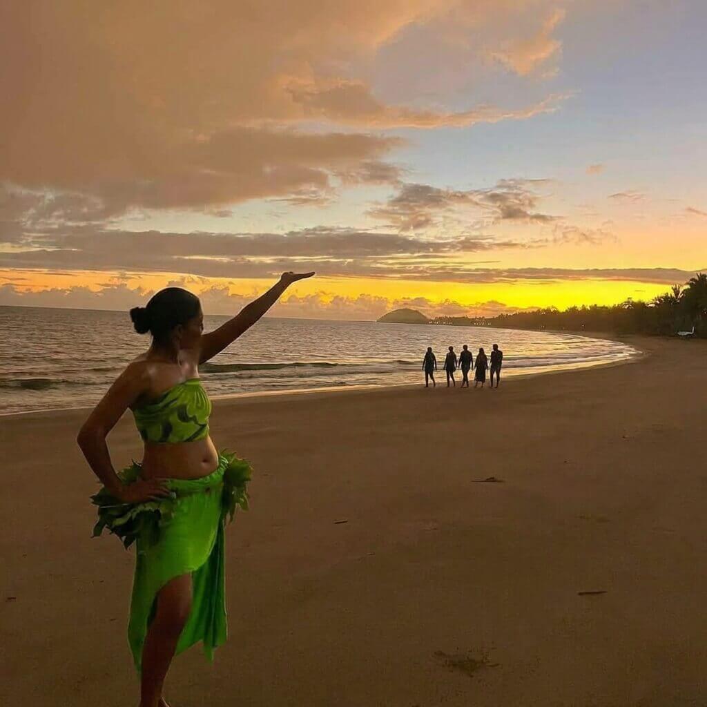A woman presents a beautiful sunset on the beach front at Uprising Resort Fiji with people walking in the background