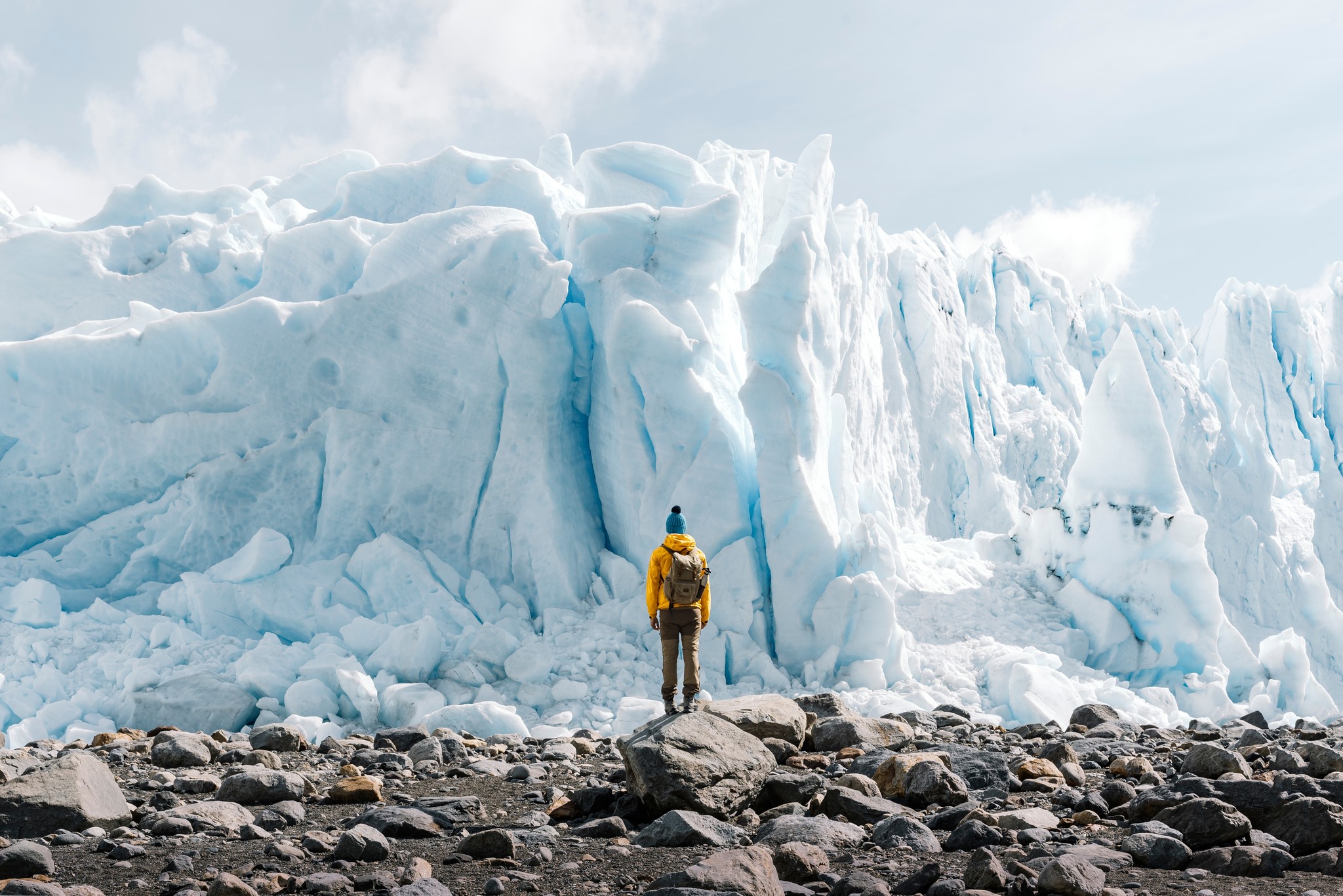 A lone hiker in a yellow jacket stands on rocky ground facing a towering wall of jagged blue ice.