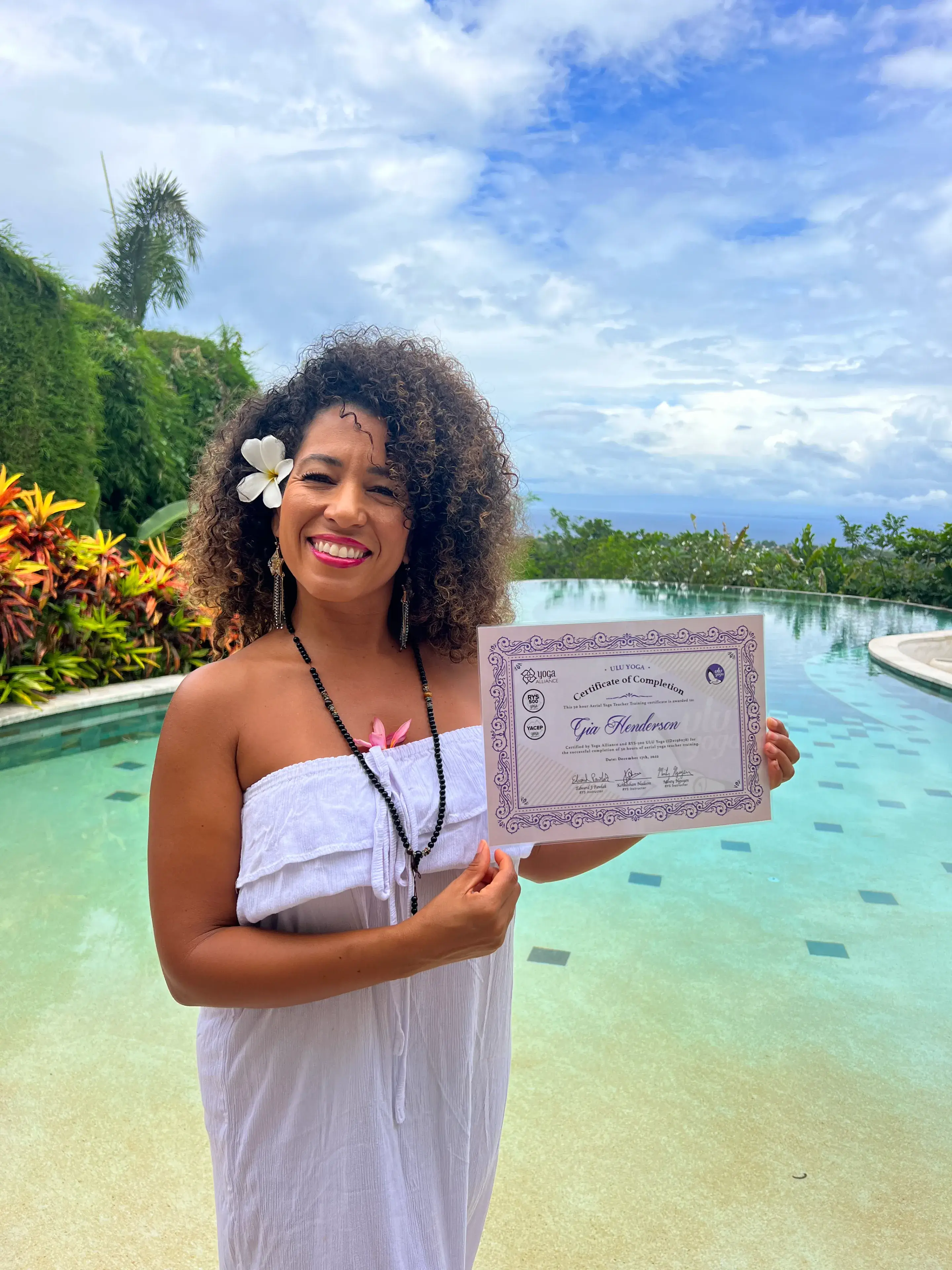 Happy graduate holding a Yoga Alliance multi-style yoga teacher training certificate by the infinity pool at Ulu Yoga Bali