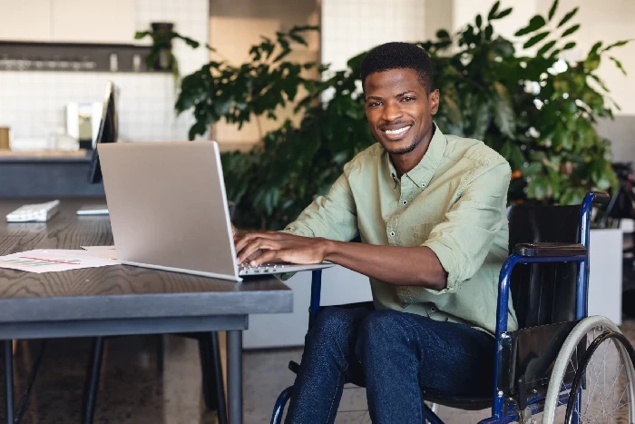 A imagem mostra um homem sentado em uma cadeira de rodas, trabalhando em um laptop e sorrindo para a câmera. Ele está em um ambiente de escritório moderno, com plantas ao fundo, o que contribui para um ambiente acolhedor e profissional. O homem veste uma camisa verde clara e parece estar envolvido em alguma atividade produtiva no computador. A cena transmite uma sensação de inclusão e acessibilidade no ambiente de trabalho, destacando uma atmosfera positiva e de integração.