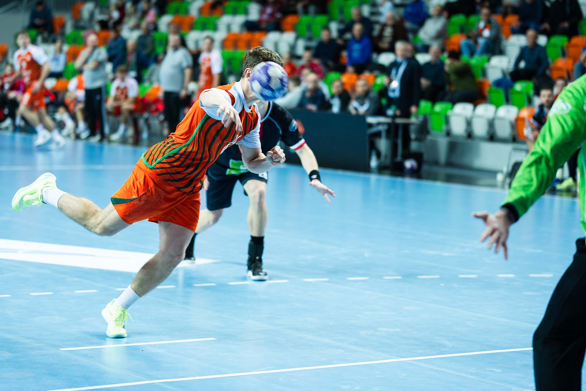 Male handball player in an orange striped jersey leaps and throws the ball toward the goal during an indoor match before a partial crowd.