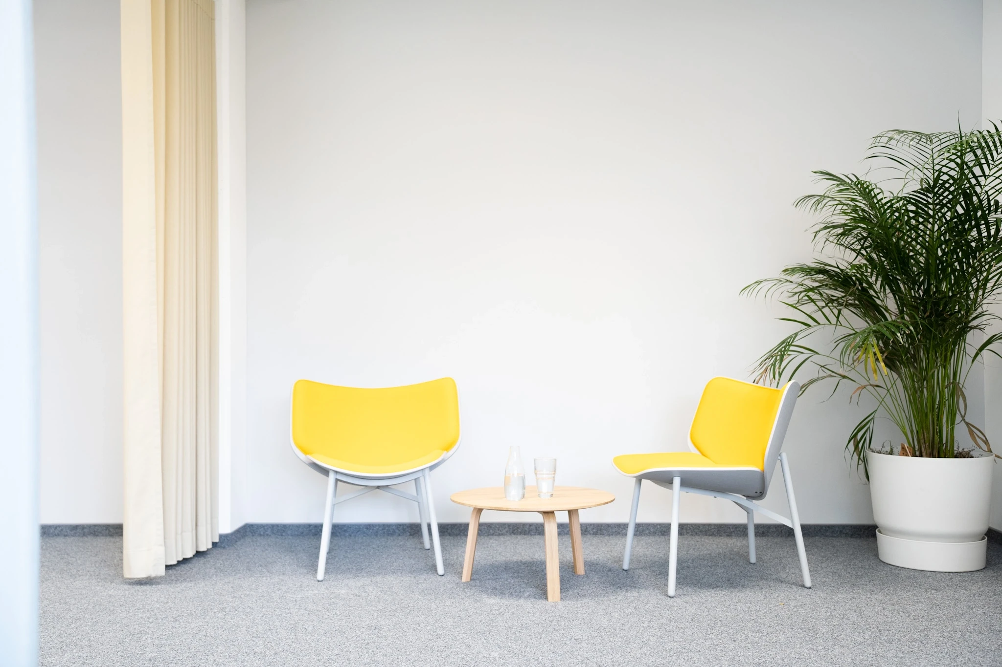 Two yellow chairs with pale grey frames around a small round wooden coffee table at NFON, against a white wall with a large potted palm and a cream curtain.