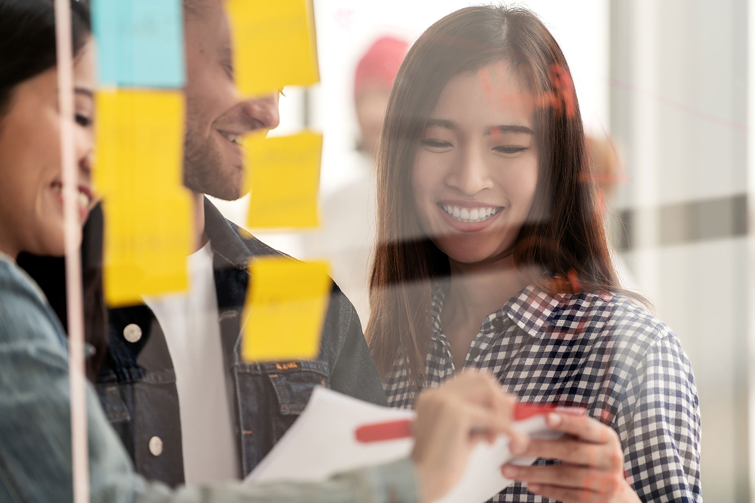 Three colleagues smiling and collaborating on a glass wall covered in yellow sticky notes.