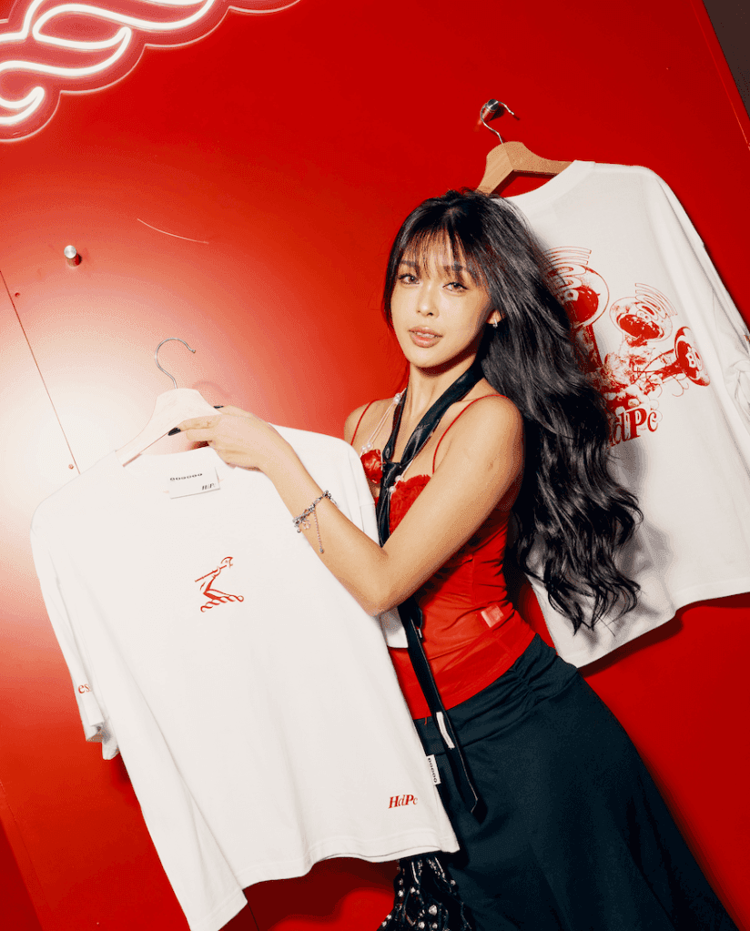 A person holding a white t-shirt with a graphic design, standing in front of a red backdrop with decorative lighting.