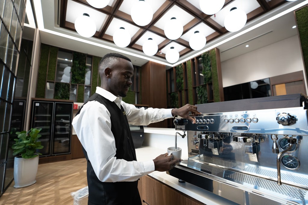 باريستا بزي رسمي يحضر القهوة باستخدام ماكينة إسبريسو احترافية في بيئة عصرية أنيقة، مما يعكس مستوى الخدمة الراقية التي تتوقعها عند زيارة أفخم مقاهي في أبوظبي.