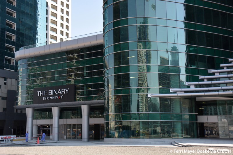 A modern cityscape featuring The Binary by Omniyat, a large building with a distinctive green glass exterior.