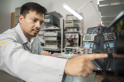 A man focuses on a screen, using his index finger to operate it, in a room filled with tech devices and wiring.