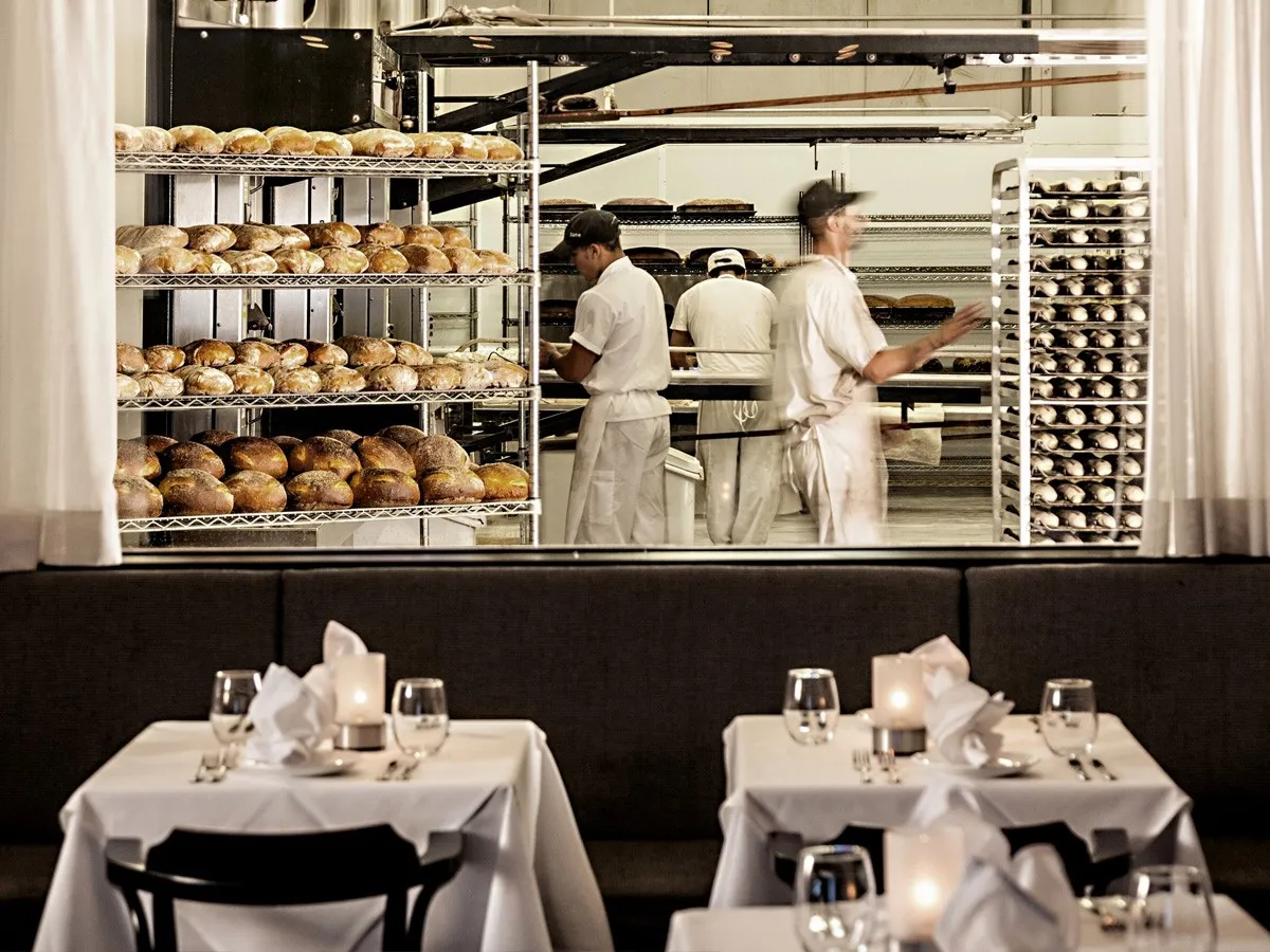 Pain d'Avignon bakers loading freshly baked bread onto shelving racks in the production kitchen, with the restaurant dining room visible below