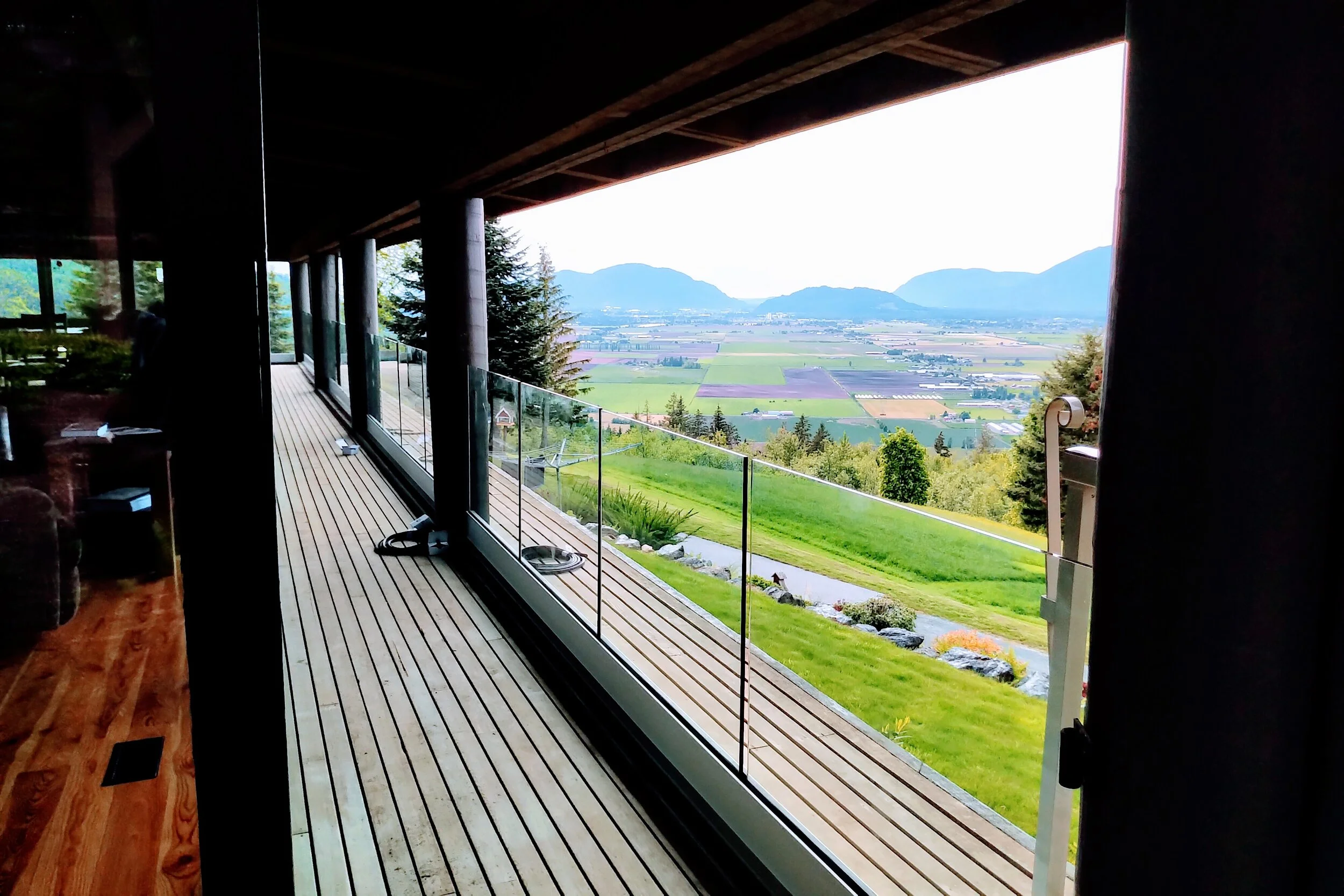 Long wooden balcony with frameless glass railing overlooking farmland and mountain views in Vancouver, designed and installed by Tenmar.