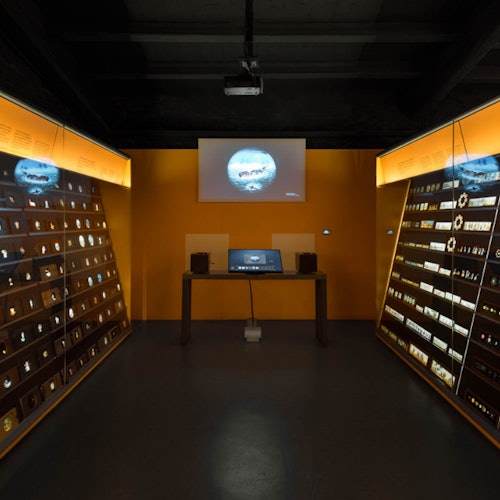 A dimly lit room with illuminated display cases on both sides, showcasing various items. A desk with a monitor and speakers is centered by a projected screen.