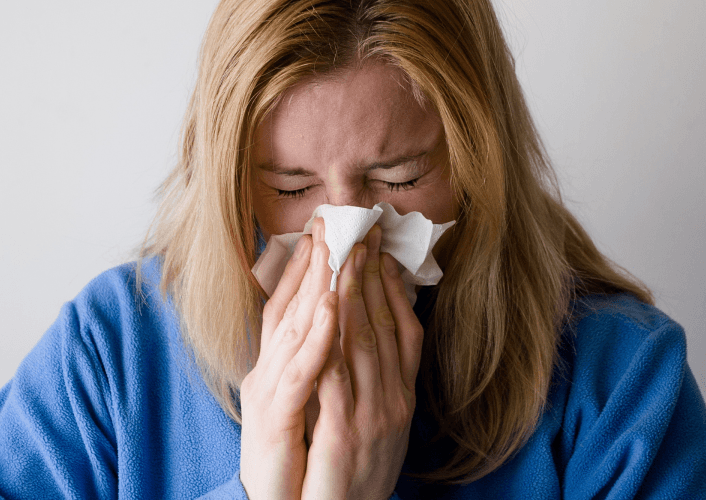 Woman sneezing into a tissue, appearing unwell against a plain background.