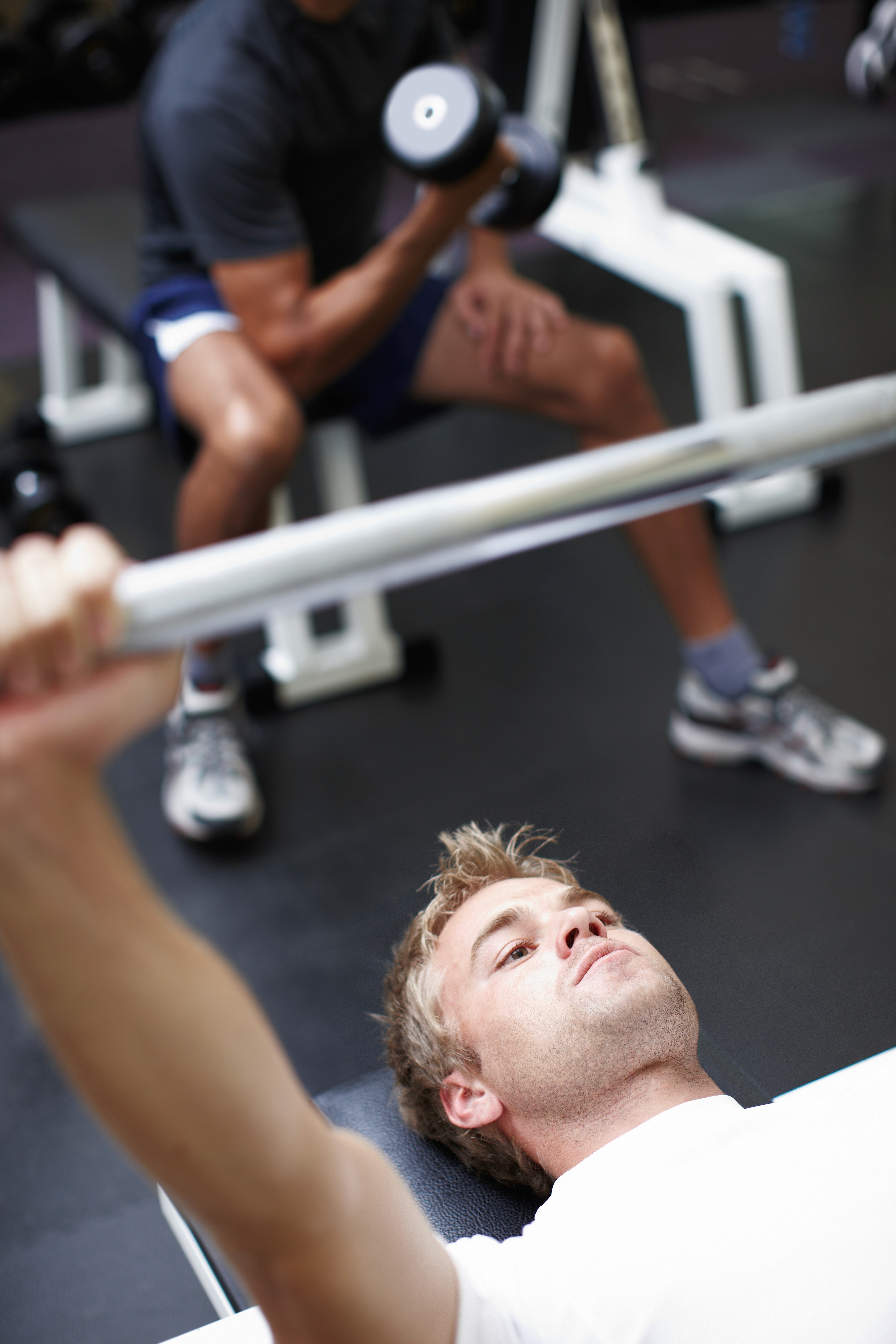 A young man lying on a bench in the gym performing a bench press exercise. He is holding a barbell, focusing on his movement. In the background, another person is seated, performing a bicep curl with a dumbbell. The background individual is wearing a dark shirt and blue shorts, but is slightly blurred, directing the focus to the man doing the bench press. The gym setting is equipped with standard weight training equipment, and the atmosphere suggests a shared training environment.