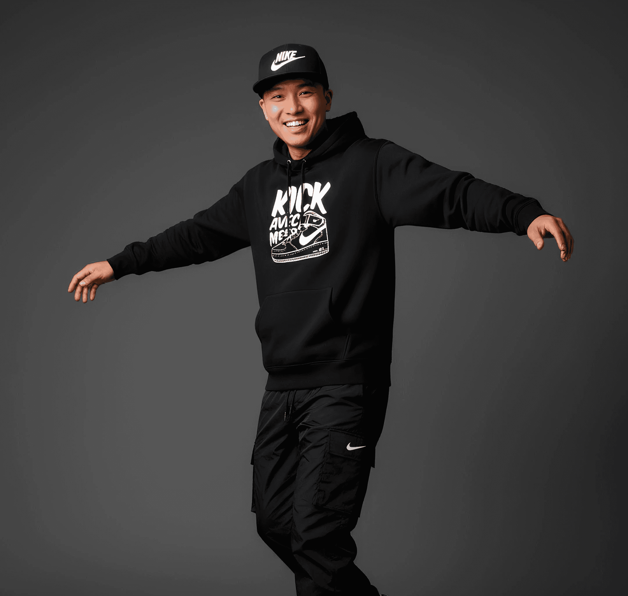 Photo de studio en pleine longueur d'un homme asiatique souriant, au style streetwear dynamique. Il porte une casquette noire 'Nike', un sweat à capuche noir avec un motif graphique blanc central ('Kick Avec Moi' ou similaire), un pantalon cargo noir à fermeture éclair avec un petit logo Nike, et des sneakers blanches et violettes de type Jordan 4. Il est en mouvement ou en pose ludique, les bras écartés. Le fond est un uni gris anthracite sombre.