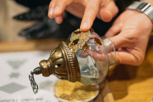 A person crafting a Turkish mosaic lamp, carefully applying multicolored beads to the ornament. The background shows a workshop setting with tools hanging on the wall.