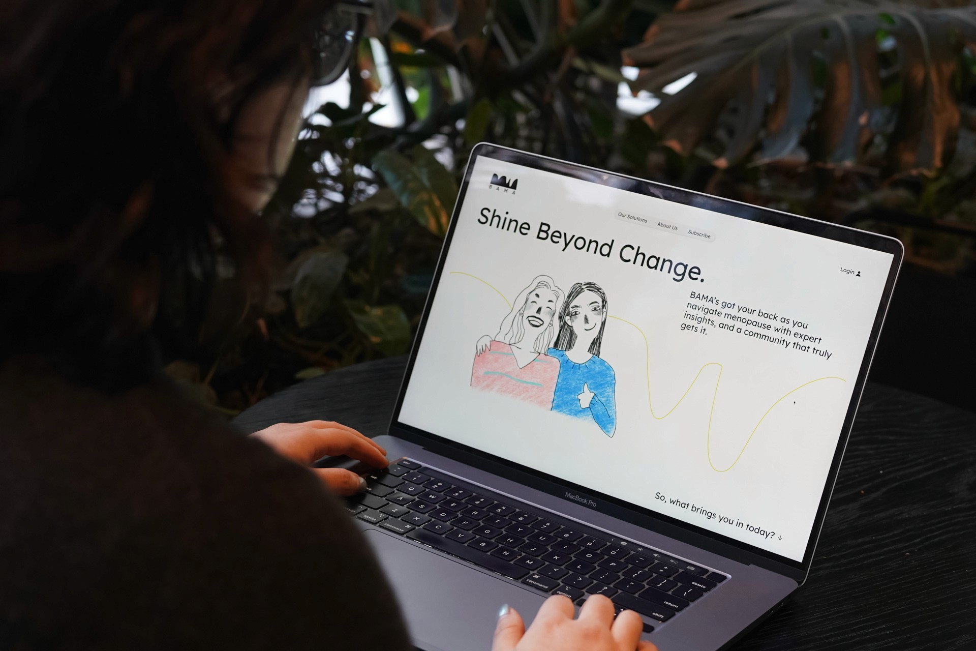 A person with dark hair is using a silver laptop on a dark surface. The laptop screen displays a graphic with the text "Shine Beyond Change." Below the graphic, a text description reads: “BAMA’s got you back as you navigate menopause with expert insights, and a community that truly gets it.” A yellow line graph is visible on the screen. The person’s hands are on the keyboard. The background is out of focus, showing green foliage.