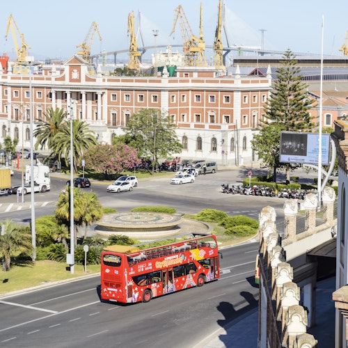 A red double-decker tour bus on a city street with palm trees, a large brick building, and industrial cranes in the background.