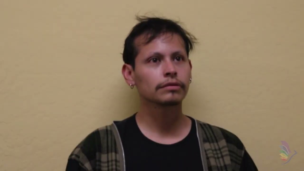 A person with short, dark hair and a mustache is wearing a plaid jacket over a black shirt, standing against a plain yellow wall.