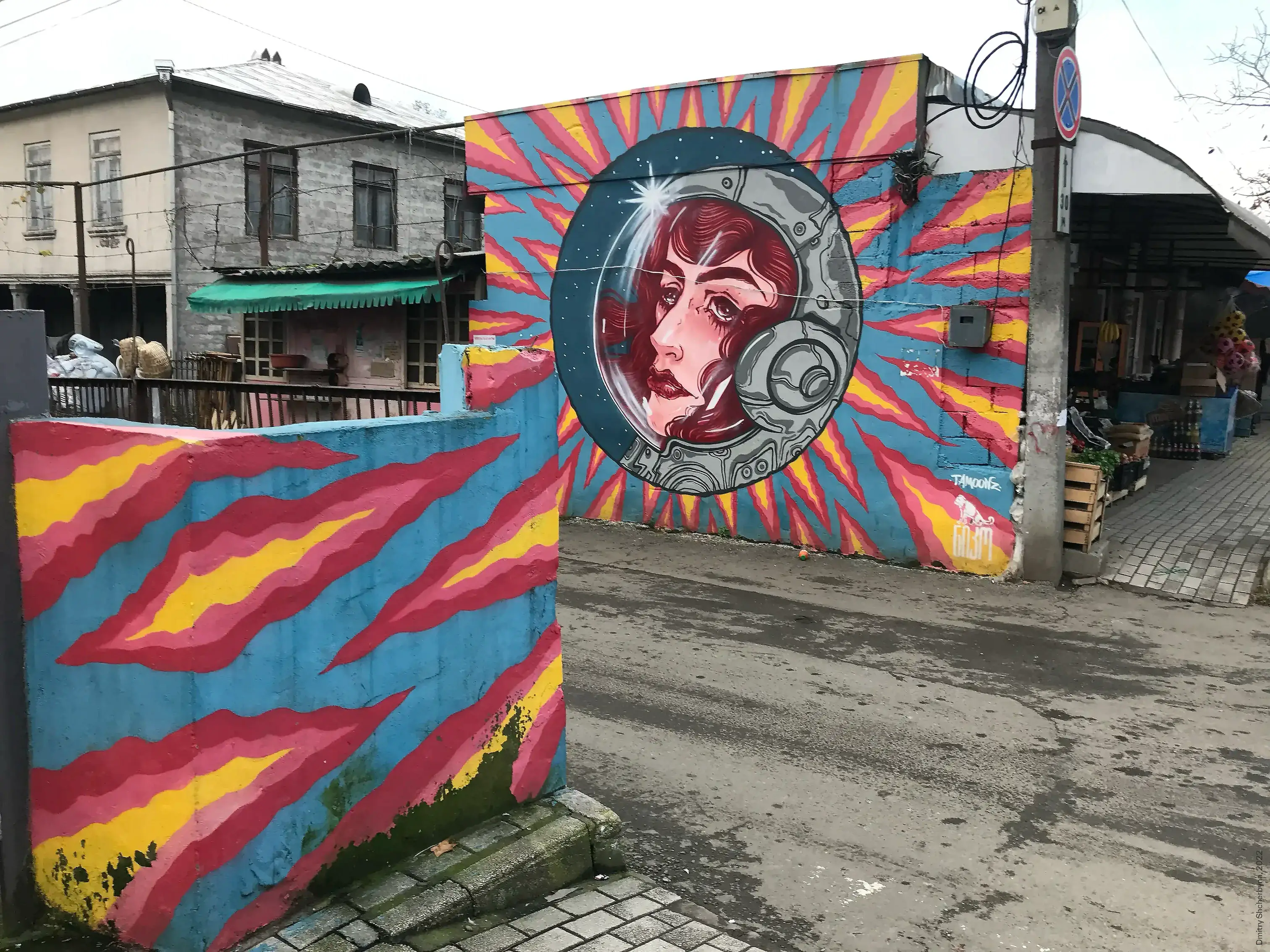 Colourful street-level mural of a woman's face inside a retro space helmet, set within a circular frame surrounded by radiating pink, yellow, and blue stripes.