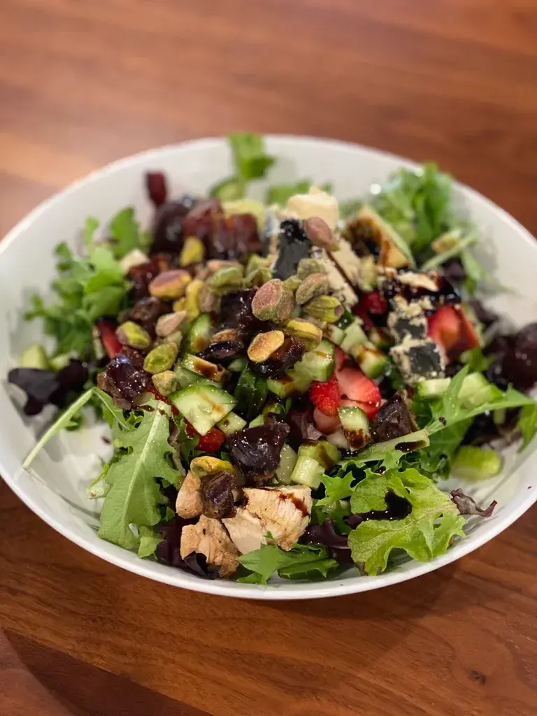 A white bowl filled with a fresh salad of mixed greens, grilled chicken, sliced strawberries, cucumbers, grapes, pistachios, and cheese on a wooden table.
