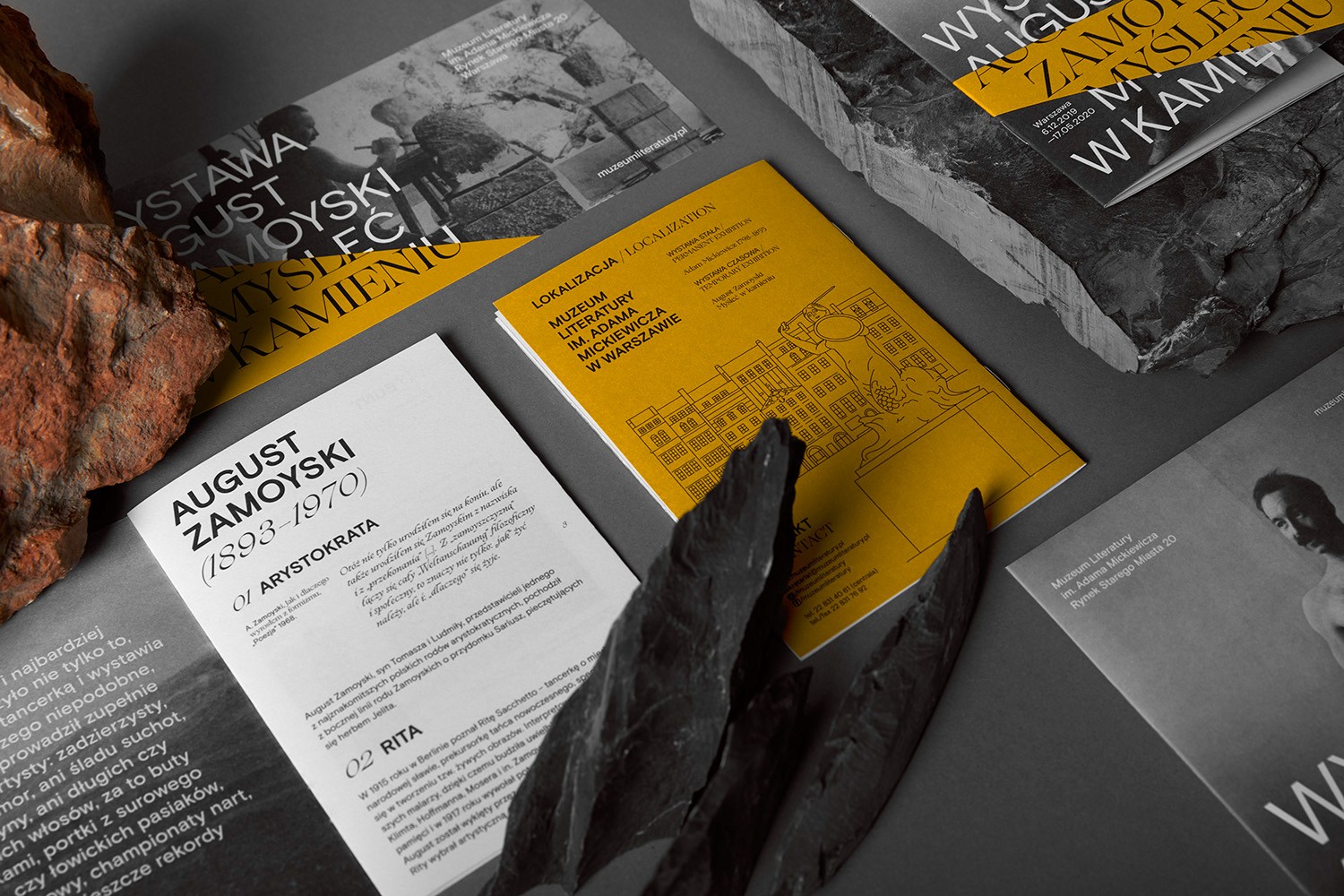 An overhead flat-lay of the exhibition's printed materials, including a yellow location map and biographical brochures, surrounded by raw stone fragments. This showcases a cohesive brand ecosystem and meticulous attention to typographic detail in museum communication.