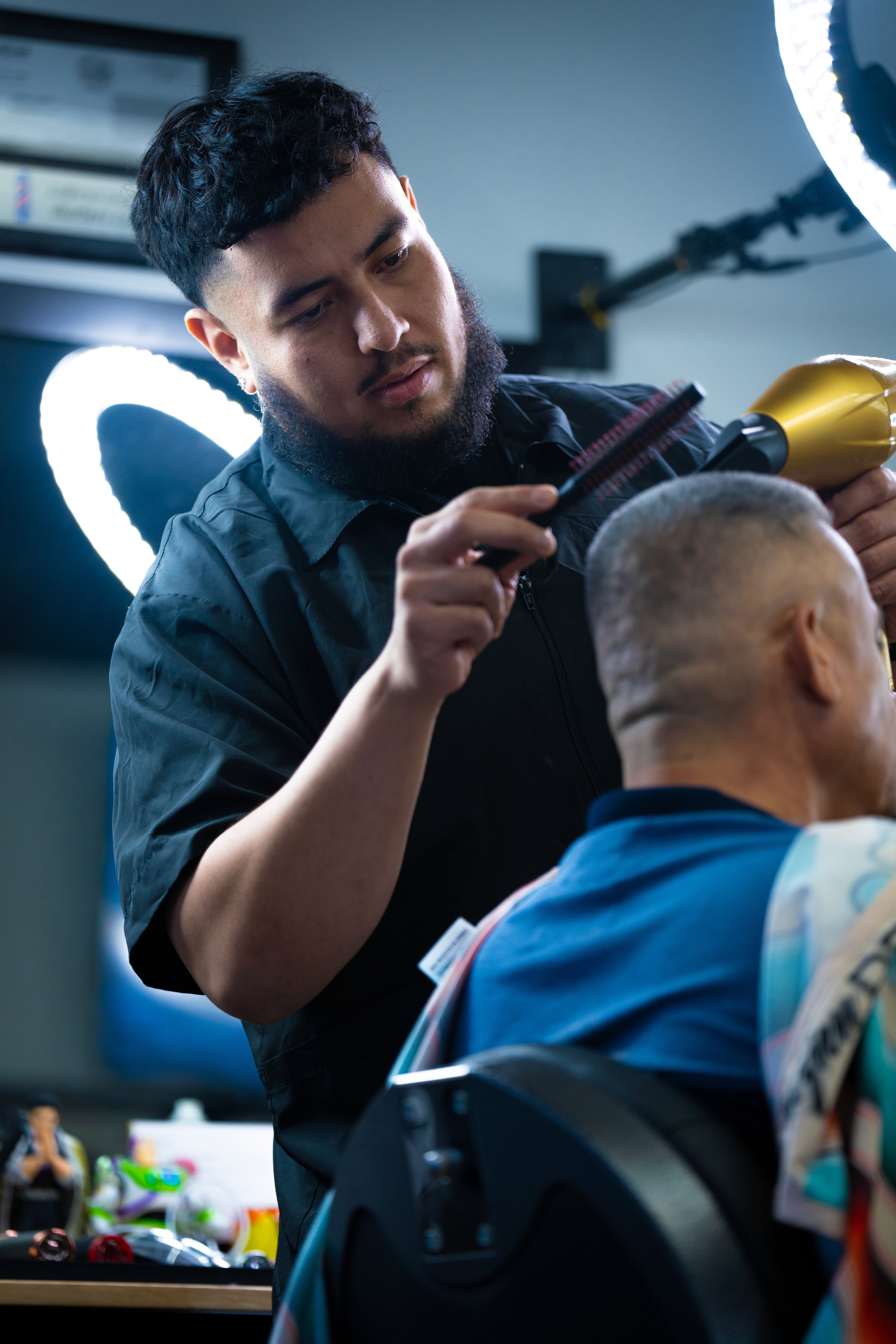 A barber with a black shirt stands and uses a blow dryer and a comb to shape a clients hair. 