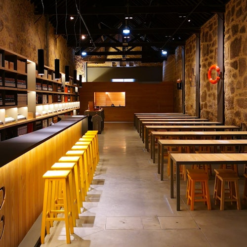 Bar moderno com bancos amarelos, longas mesas de madeira e paredes de pedra. Prateleiras de garrafas e um balcão iluminado são visíveis.