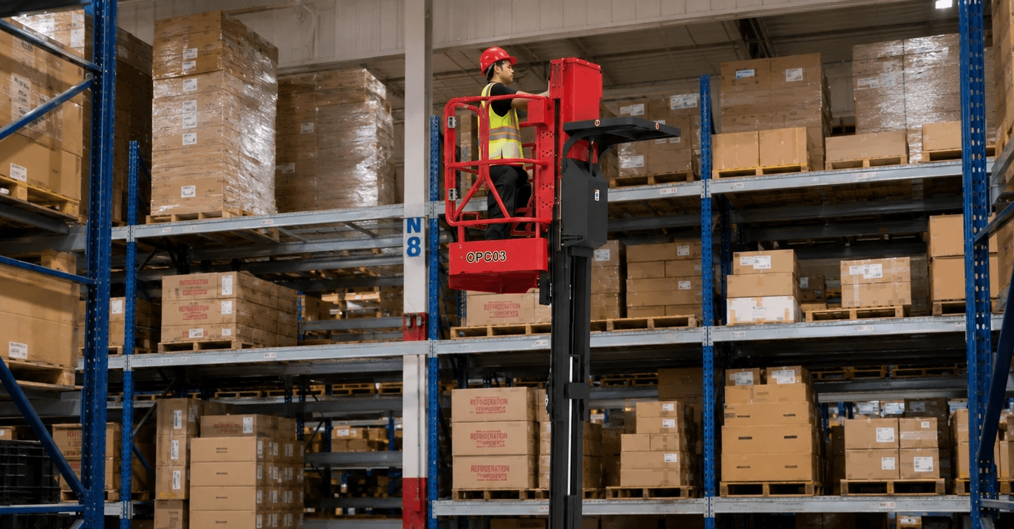 A red and black order picker forklift working alongside warehouse shelving, with an operator standing on the lift platform to retrieve and store items.