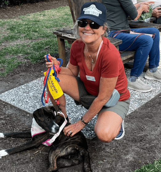 A woman kneels beside a black dog, smiling while holding colorful leashes in a grassy outdoor setting.
