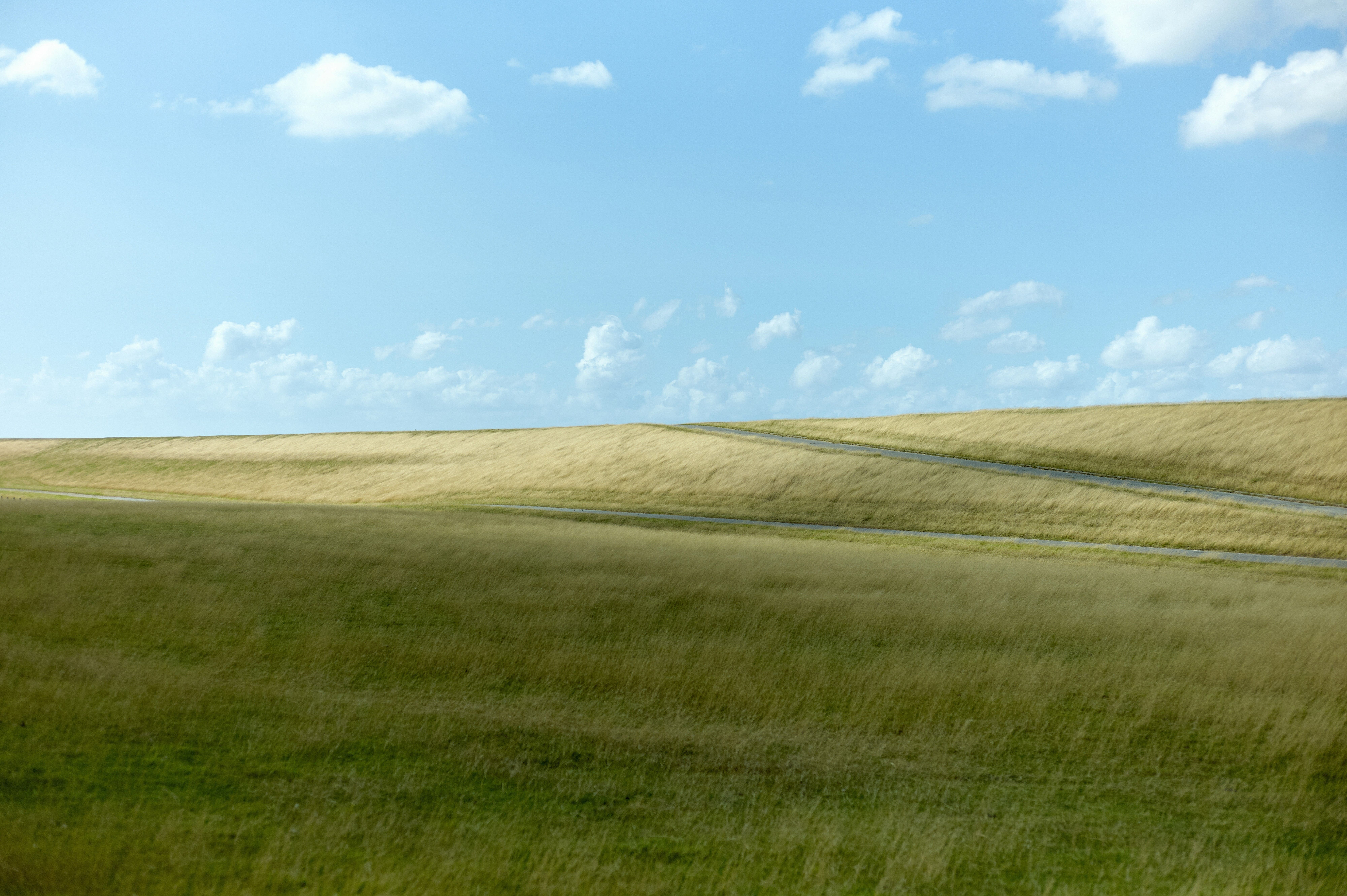Wide landscape of rolling green and yellow grassy hills with a narrow asphalt road under a blue sky.