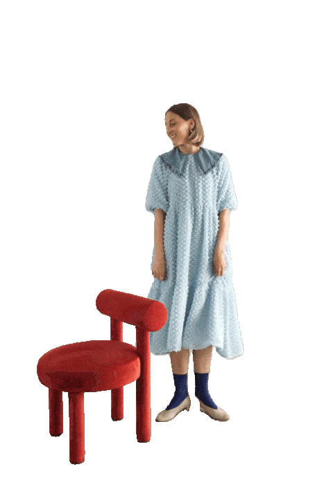 A woman in a blue gown dancing next to a red, modern chair with rounded edges, smiling and looking to the side.