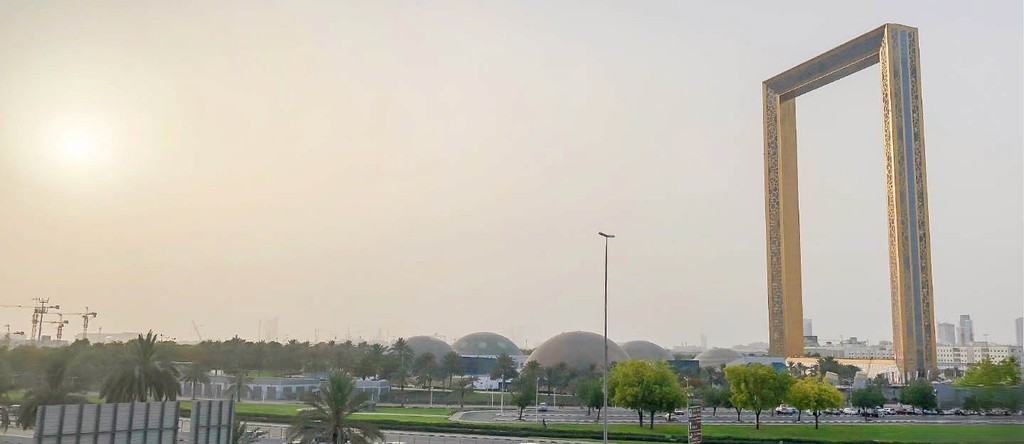 Dubai Frame landmark in Al Jaddaf with golden design, domes, trees, and hazy sky at sunset.