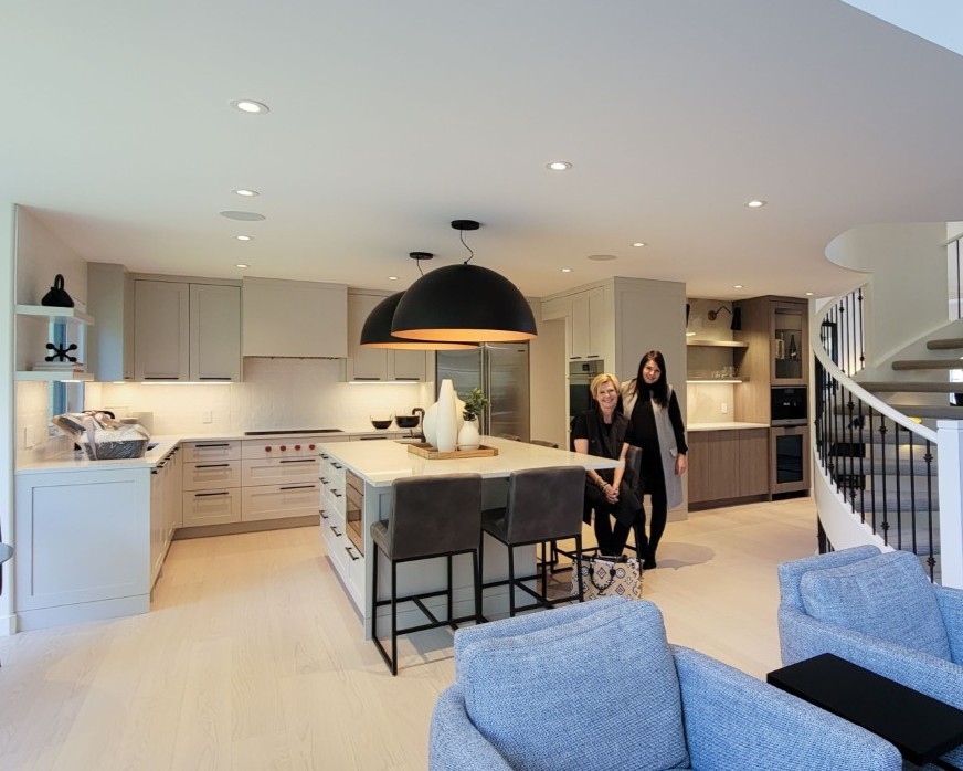 A green/grey kitchen with dark oversized dome lights over the island. Two fair skinned women stand beside the island smiling at the camera.