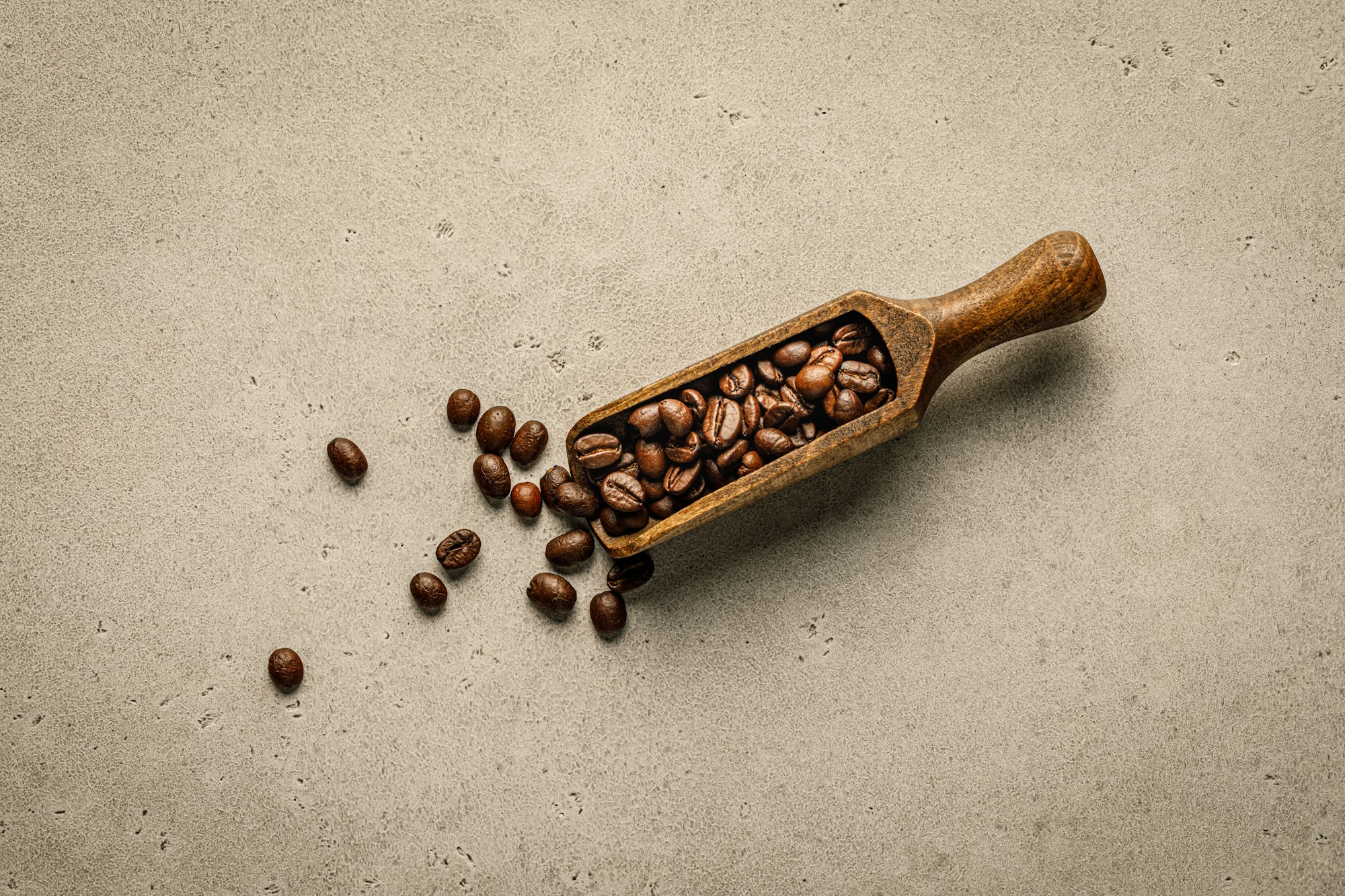 Wooden scoop of roasted coffee beans with loose beans on a stone surface