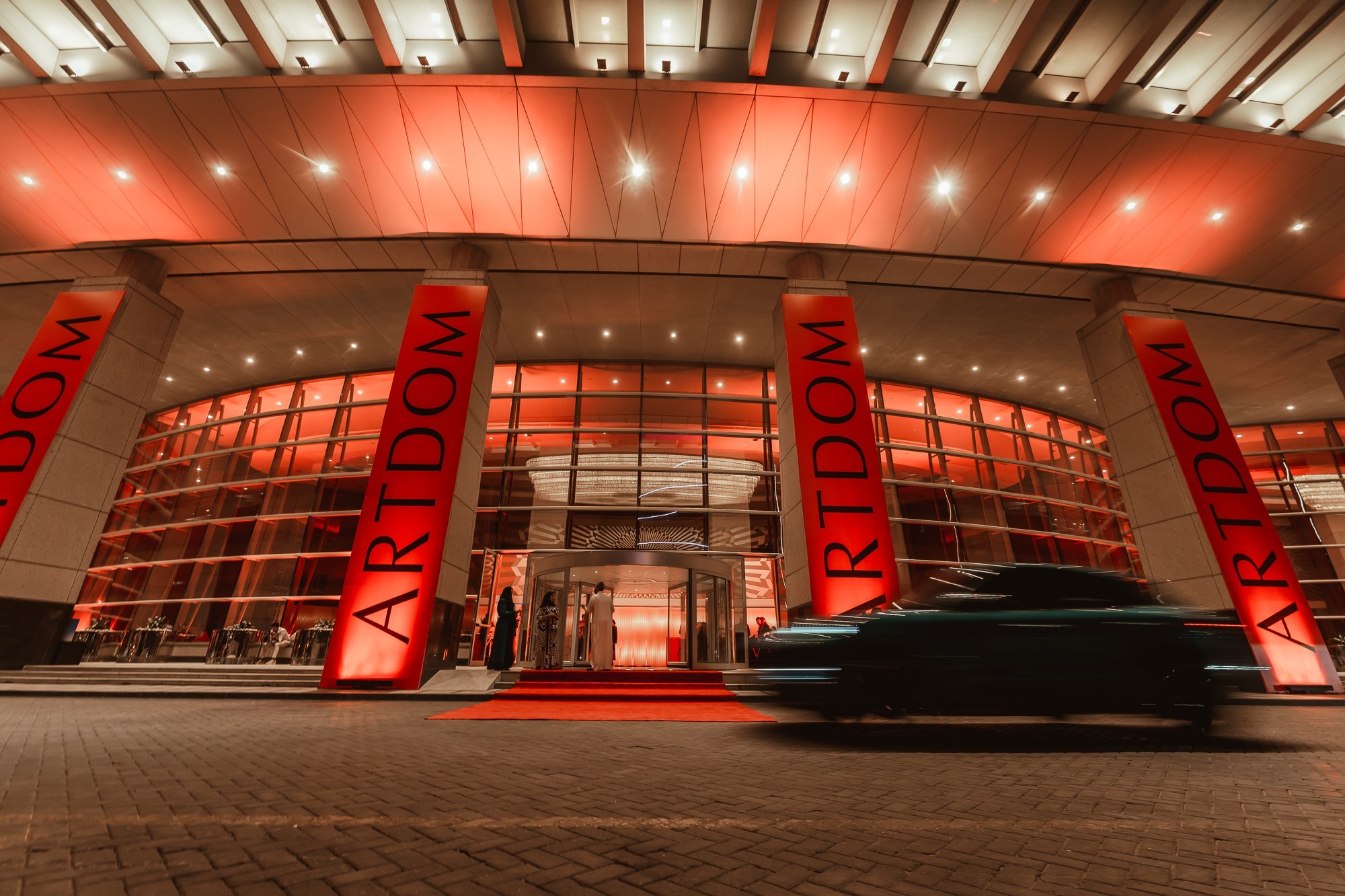 Hotel entrance lit in red with tall ARTDOM banners on its columns and a car passing in motion blur