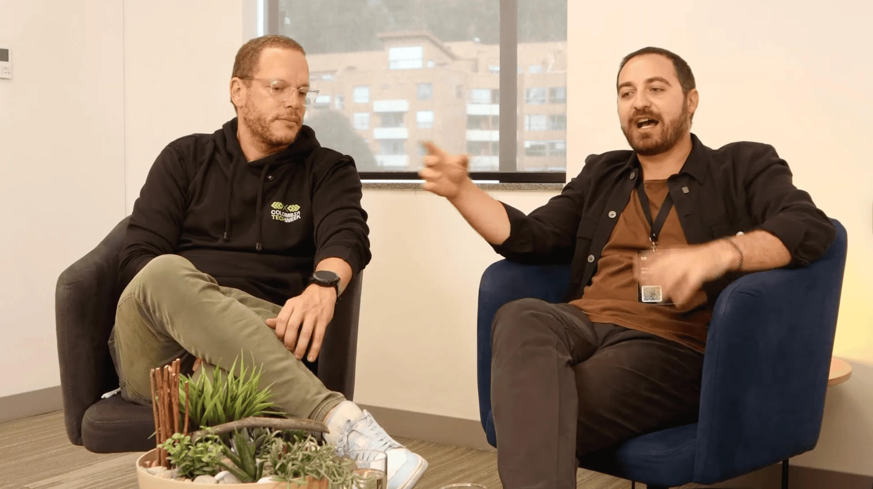 Dos personas sentadas en sillones conversando de forma relajada durante una charla, en un espacio interior con plantas decorativas y luz natural al fondo.