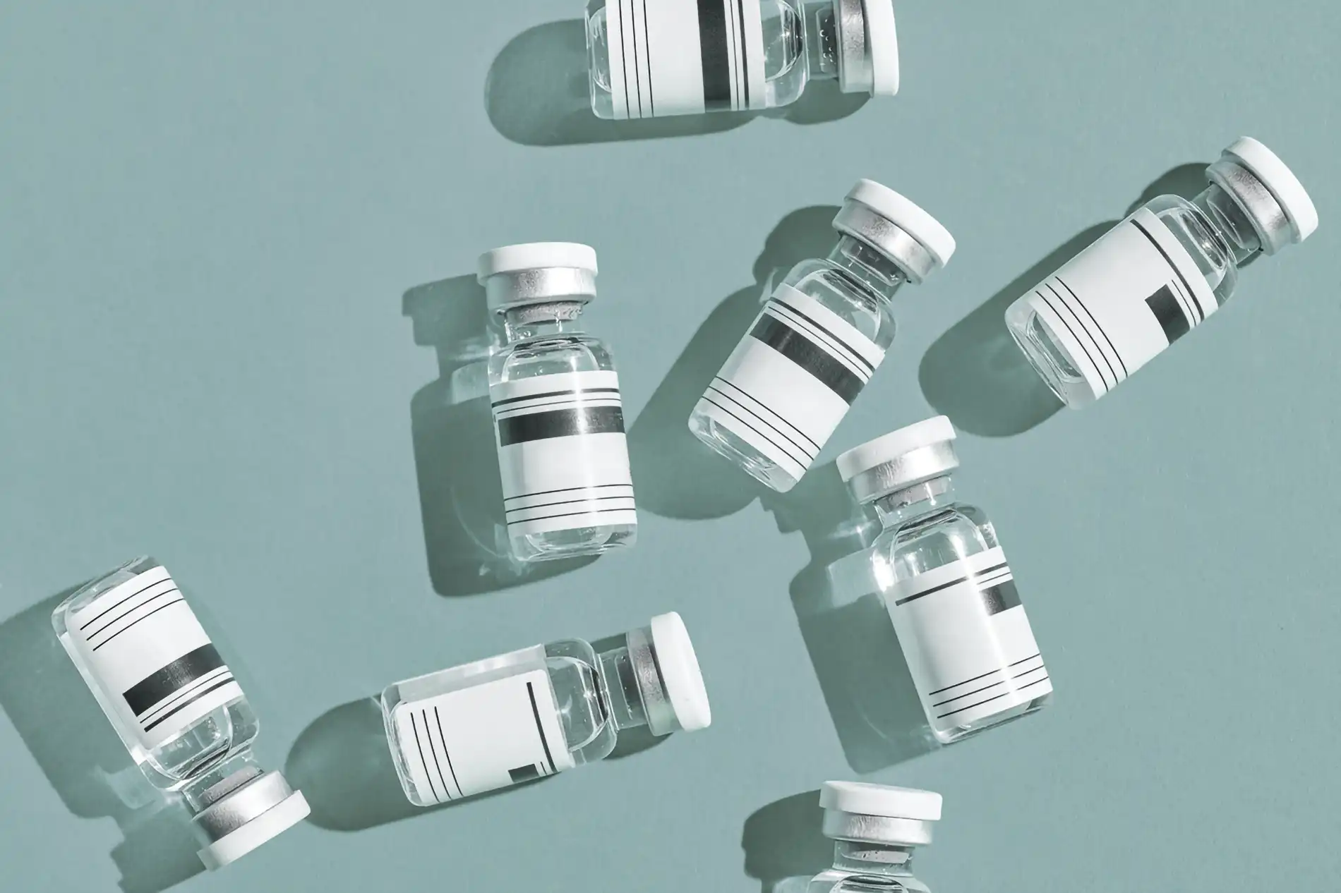 Several glass vials with white labels arranged on a muted blue-green background, casting shadows in direct sunlight.