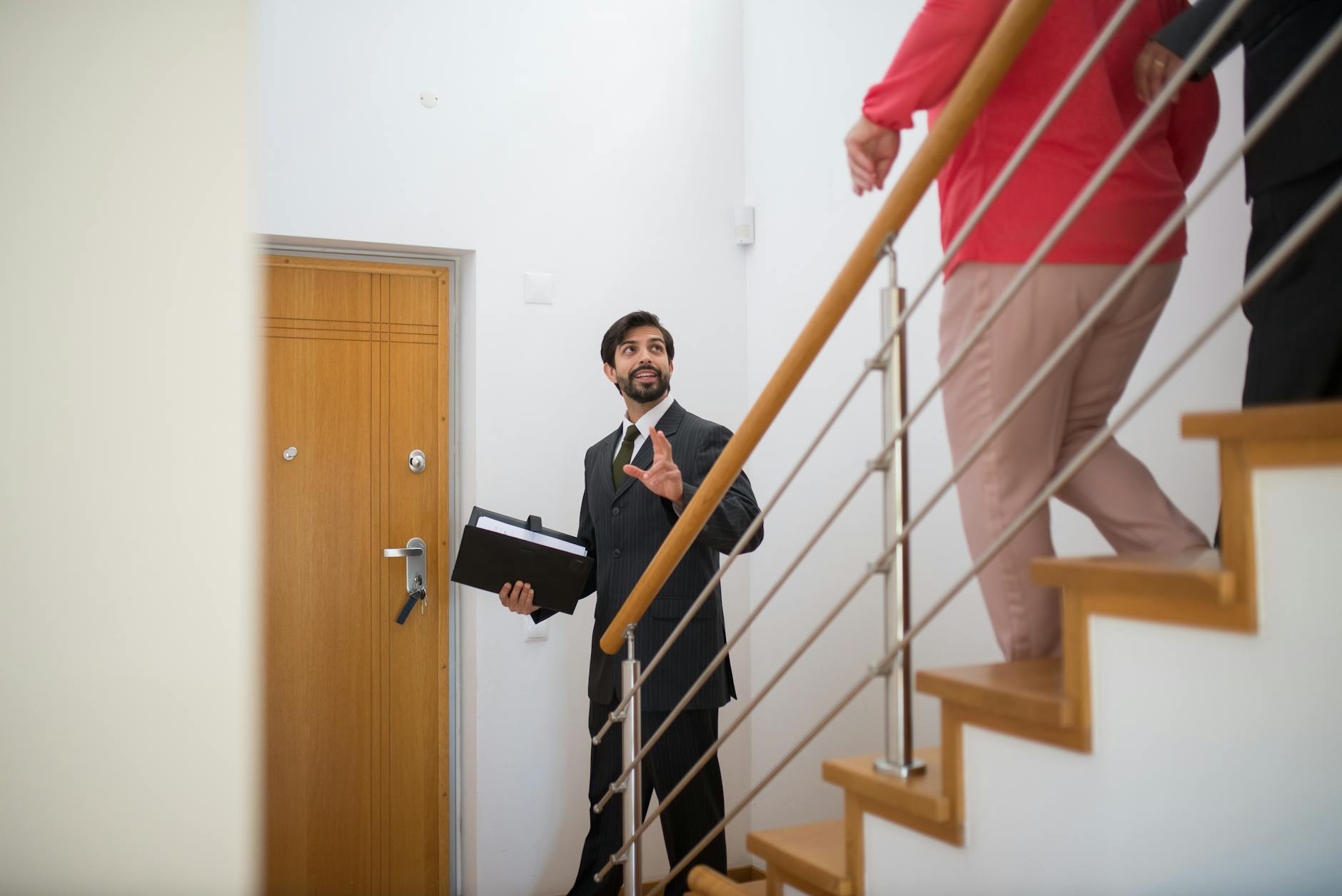 Suited letting agent holding a folder gesturing to clients ascending a wooden staircase inside a property